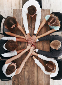 Top view of meeting for the business team, brainstorming on the new project. Co-workers sitting at the brown wooden desk. Colleagues putting hands together while working on project indoors. Unity concept